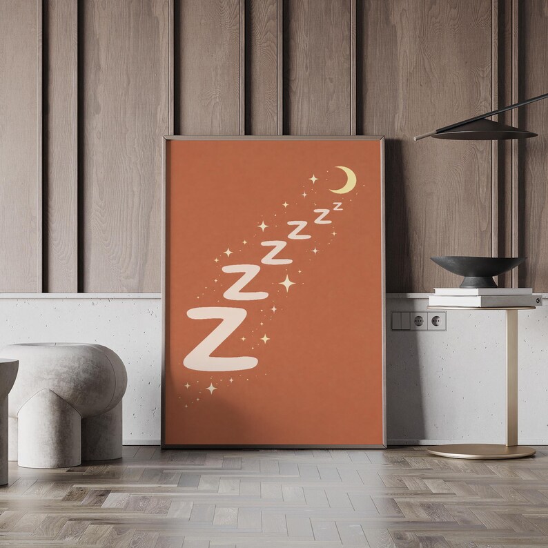 May include: A framed art print with a terracotta background features the word "Zzz" repeated in a diagonal line, accompanied by stars and a crescent moon. The letters are off-white, creating a calming, minimalist design. The artwork is displayed against a wooden wall.