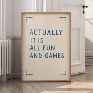Pode incluir: Uma impressão emoldurada com o texto "ACTUALLY IT IS ALL FUN AND GAMES" em azul. A impressão tem um fundo creme e uma moldura de madeira. Acentos azuis decorativos estão em cada canto.