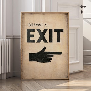 May include: A framed sign featuring the words "DRAMATIC EXIT" in black, with a black hand pointing right. The sign has a distressed, vintage aesthetic. The background is a neutral colour, and the frame is wooden.