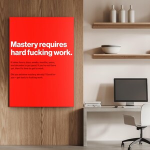Puede incluir: Un lienzo rectangular rojo con texto blanco que dice "Mastery requires hard fucking work." Texto adicional debajo detalla el esfuerzo necesario para lograr el dominio. El lienzo cuelga de una pared de madera.