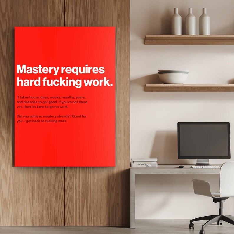 Puede incluir: Un lienzo rectangular rojo con texto blanco que dice "Mastery requires hard fucking work." Texto adicional debajo detalla el esfuerzo necesario para lograr el dominio. El lienzo cuelga de una pared de madera.