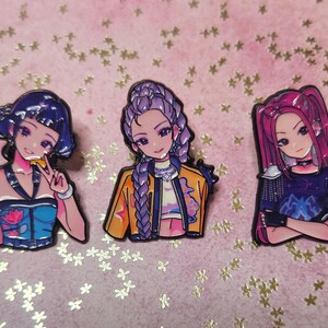 May include: Three anime-style enamel pins depicting female characters with varied hairstyles and clothing. The pins have a glossy finish and are set against a pink background with gold star-shaped confetti.