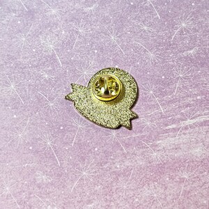 May include: Gold-colored enamel pin with a heart shape and a banner. The back of the pin has a safety clasp.