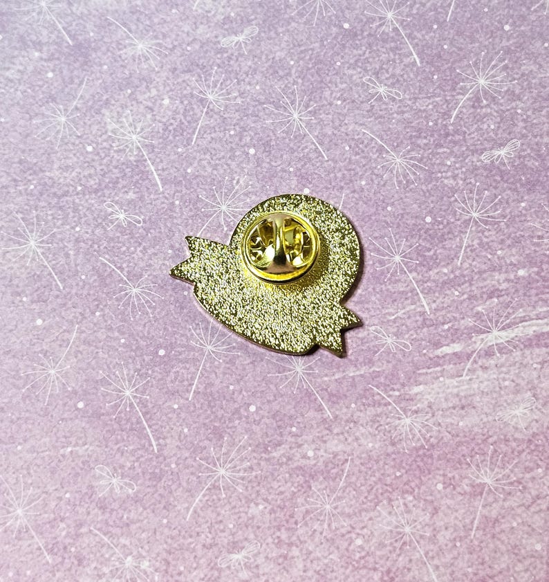 May include: Gold-colored enamel pin with a heart shape and a banner. The back of the pin has a safety clasp.