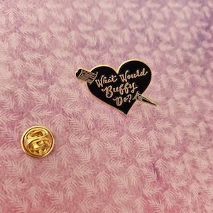 May include: A gold-coloured pin and a black heart-shaped enamel pin with the text "What Would Buffy Do?" in gold lettering. The heart is pierced by a gold arrow. The background is a pink and white floral pattern.