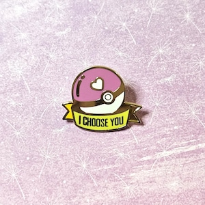 May include: A gold-colored enamel pin featuring a pink and white Pokeball with a small heart on top. The pin has a yellow banner that reads "I CHOOSE YOU".