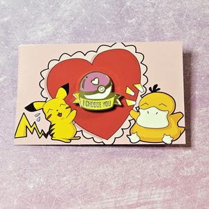 May include: A pink and gold Pokemon pin with a Pokeball and the text "I Choose You". The pin is on a pink card with a red heart and cartoon images of Pikachu and Psyduck.