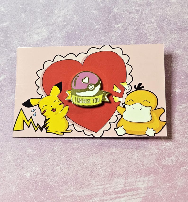 May include: A pink and gold Pokemon pin with a Pokeball and the text "I Choose You". The pin is on a pink card with a red heart and cartoon images of Pikachu and Psyduck.