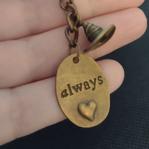 May include: Bronze-coloured keyring with a round ring, chain, and charm. The charm features an oval tag with the word "always" and a heart. A small hat charm hangs from the chain. The keyring is held in a hand.