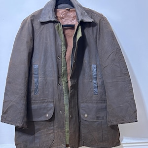 May include: A brown waxed cotton jacket with a leather collar and accents, hanging on a hanger. The jacket features multiple pockets, a button-up front, and a green lining. A classic outerwear garment.