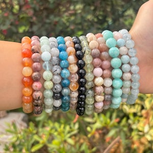 May include: A selection of beaded bracelets in a range of colours, including orange, pink, green, blue, brown, black, and light blue. The bracelets are made of round gemstone beads and are shown on a wrist.