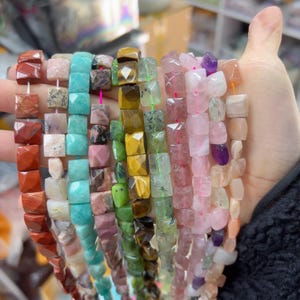 Peut inclure: Plusieurs brins de perles de pierres précieuses sont tenus dans une main. Les perles sont taillées en forme carrée et enfilées sur des cordons colorés. Les pierres précieuses sont de couleurs variées, dont le rouge, le rose, le vert et le bleu. Les perles mesurent environ 1,3 cm.