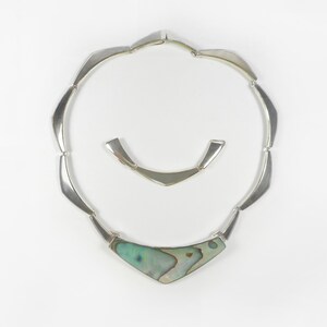 May include: A silver necklace with a geometric design and a large abalone shell pendant. The necklace is made of interlocking silver pieces that form a V-shape.