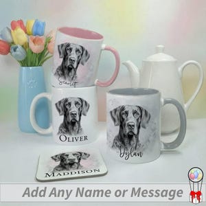 May include: A collection of white ceramic mugs featuring dog portraits and custom names. One mug has a pink interior and handle, another has a grey interior and handle. A matching coaster is also visible. A blue vase with flowers and a white teapot are in the background.