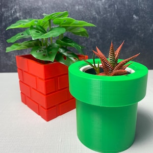 May include: Two decorative planters. One is a red brick design, the other a green pipe. The red planter holds a green leafy plant, and the green planter has a small succulent with red-tipped leaves.