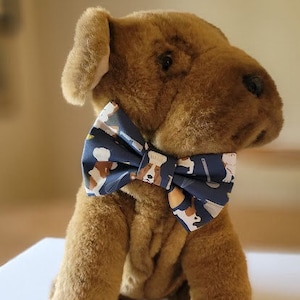 Pode incluir: Um cão de pelúcia marrom usando uma gravata borboleta azul com uma estampa de cachorro branco e marrom. A gravata borboleta tem um fundo branco com uma borda azul.