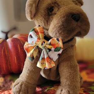 May include: A brown plush dog wearing a bow tie with a autumn-themed print. The bow tie has orange, yellow, and white colours with a pattern of pumpkins, leaves, and acorns. The dog is sitting on a brown and orange patterned fabric.