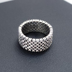 May include: A silver ring with a repeating pattern of small, clear stones. The ring has a wide band and a textured surface, creating a sparkling effect. The ring is set against a dark background.