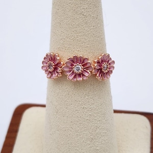 May include: A rose gold ring with three pink enamel daisy flowers with clear cubic zirconia stones in the center of each flower.