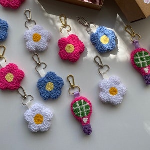 May include: A collection of handmade keychains in the shape of flowers and a tennis racket. The flower keychains are in pink, white, and blue with yellow centers. The tennis racket keychain is pink and green. All keychains have gold-colored clasps.