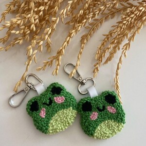 May include: Two handmade green frog keychains with black eyes and pink cheeks. The keychains are made of a textured yarn and have silver-colored clasps and white ribbon loops. The background features dried wheat stalks.