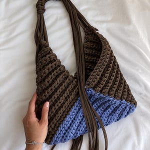 May include: A crochet hobo bag featuring a dark brown upper section and a light blue lower section. The bag is constructed with a ribbed stitch pattern and includes a long, braided dark brown strap with several dangling fabric fringe strands.