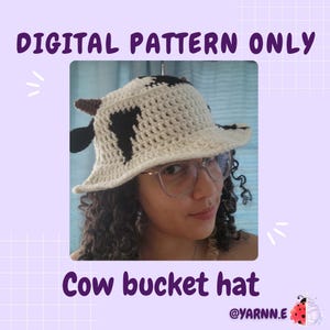 Puede incluir: Un sombrero de pescador de ganchillo con base blanca, manchas negras de vaca y pequeños cuernos marrones. El texto DIGITAL PATTERN ONLY y Cow bucket hat aparecen en la imagen, junto con el nombre @YARNN.E y un icono de mariquita.