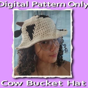 May include: A crocheted cow bucket hat with black and white spots. The hat is being worn by a person with curly hair.