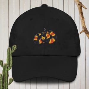 Puede incluir: Gorra de béisbol negra con una imagen bordada de una serpiente roja, amarilla y negra.