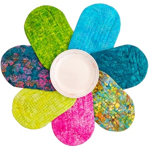 Könnte beinhalten: Ein Set aus sieben bunten, gesteppten Platzsets, die in Form einer Blume um einen weißen Teller angeordnet sind. Die Platzsets sind in Grün-, Blau-, Türkis-, Gelb- und Rosatönen gehalten.