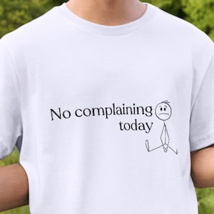 May include: White t-shirt with the text "No complaining today" and a stick figure illustration. The shirt is short-sleeved and appears to be made of a soft material. The background is blurred, with green foliage.