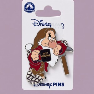 Op de afbeelding: Een Disney Pins emaille speldje met Grumpy uit Sneeuwwitje, gekleed in een rood jasje en geruite pantoffels, met een zwarte mok met de tekst KEEP Smiling. Het speldje zit op een witte kaart met het Disneyland Paris logo aan de bovenzijde.