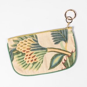 Könnte beinhalten: Eine kleine, cremefarbene Tasche mit einem grünen und gelben Blumendruck. Die Tasche hat einen goldenen Schlüsselring, der am Reißverschluss befestigt ist.