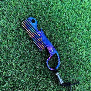 May include: A multi-tool comb featuring an iridescent, heat-anodised finish with blue, purple, and gold swirling patterns. The tool includes a row of teeth, a bottle opener notch, a hexagonal cutout, a finger ring, and a black lanyard with a metal bead.