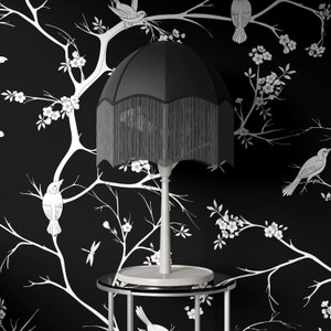 May include: A black lampshade with a fringe trim sits on a silver table with a black glass top. The lamp is on a white background with a black and white floral pattern.