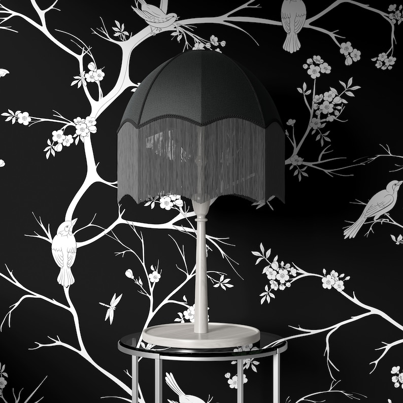 May include: A black lampshade with a fringe trim sits on a silver table with a black glass top. The lamp is on a white background with a black and white floral pattern.