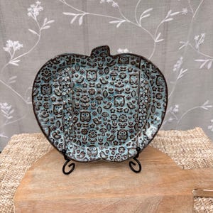 May include: A decorative ceramic plate in the shape of a pumpkin, featuring a teal and brown owl and floral pattern. The plate is displayed on a black metal stand, set against a neutral background.