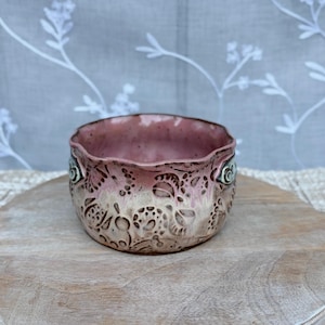 May include: A small ceramic bowl with a wavy rim, featuring a glossy pink interior and a textured exterior with a beige and brown gradient. The outer surface is decorated with intricate, embossed circular and floral patterns throughout the body.