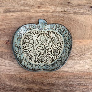 May include: A ceramic, pumpkin-shaped plate with a hand-painted sunflower design. The plate is light blue with a darker blue rim and stem. The sunflowers are white and black, with intricate details.
