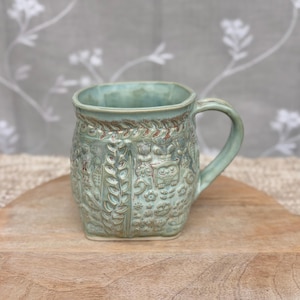 May include: A light green ceramic mug with a square-shaped body and a curved handle. The exterior surface features intricate embossed patterns of owls, flowers, and leafy vines with subtle brown highlights accentuating the textured details.