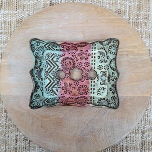 May include: A decorative ceramic light switch cover with a floral and geometric pattern. The cover features a color scheme of teal, pink, and brown. The cover has two circular holes and a star-shaped hole in the center.