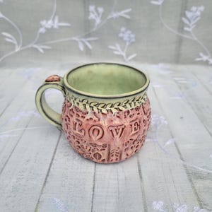May include: A handmade ceramic mug with a light green interior and a pink exterior. The word "LOVE" is embossed on the mug's exterior, surrounded by decorative patterns. The handle and rim have a cream color.