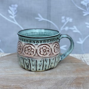 May include: A ceramic mug featuring a light blue glaze with a band of embossed floral patterns in a soft pink hue. The upper rim is decorated with a row of small circles, while the lower section displays vertical lines and dots in a textured finish.