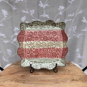May include: Square ceramic plate with a scalloped edge. The plate features a floral and geometric embossed design in shades of pink, white, and brown. The plate is displayed on a black metal stand, showcasing its decorative details.