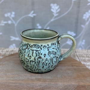 May include: A round ceramic mug with a light blue and cream glaze featuring a textured, embossed pattern of various mushrooms across the exterior surface. The mug has a smooth, curved handle and a dark, defined rim around the top opening.