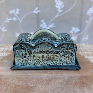 May include: A rectangular ceramic butter dish with a lid featuring a light blue and dark blue glaze. The surface is embossed with a repeating mushroom pattern and the word butter is clearly stamped in the center. It has a curved handle.