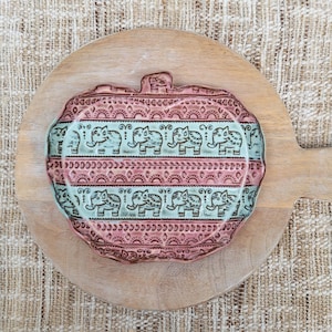 May include: A decorative, pink and teal ceramic serving platter with an elephant motif. The platter is placed on a round wooden cutting board. The elephant design is repeated in rows across the platter's surface.