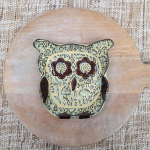May include: A ceramic owl-shaped dish with a light yellow background and dark brown accents. The dish is decorated with a pattern of mushrooms and other small shapes. The dish is sitting on a wooden cutting board.
