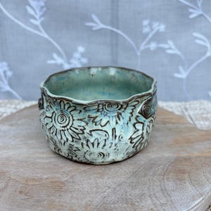 May include: A round ceramic bowl with a light blue and turquoise glaze featuring a dark brown embossed sunflower pattern. The bowl has a wavy, uneven rim and a smooth interior surface, showcasing a handcrafted aesthetic with visible texture.