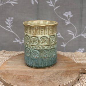 May include: A cylindrical ceramic vessel featuring a scalloped rim and a gradient glaze transitioning from beige at the top to teal at the base. The exterior is decorated with embossed vertical stripes, dotted lines, and repeating floral motifs.